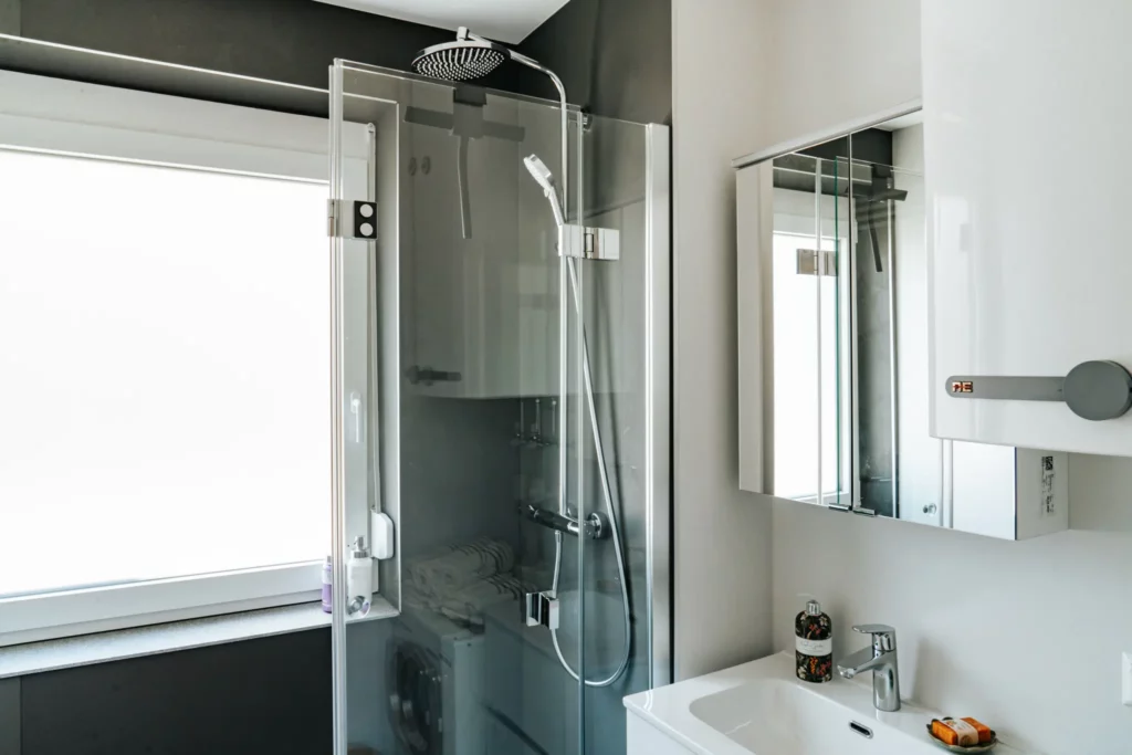 Ein modernes Badezimmer im Appartement Grand Prix in Zeltweg, ausgestattet mit einer geräumigen Glasdusche mit Regenduschkopf, einem Waschbecken mit integriertem Spiegel und stilvollen Details. Das helle Tageslicht, das durch das große Fenster fällt, schafft eine angenehme Atmosphäre und ergänzt die hochwertige Einrichtung perfekt. Ideal für Gäste, die nach einem langen Tag Entspannung suchen, sei es nach einem Besuch am Red Bull Ring oder einem Erkundungstag in der steirischen Umgebung.