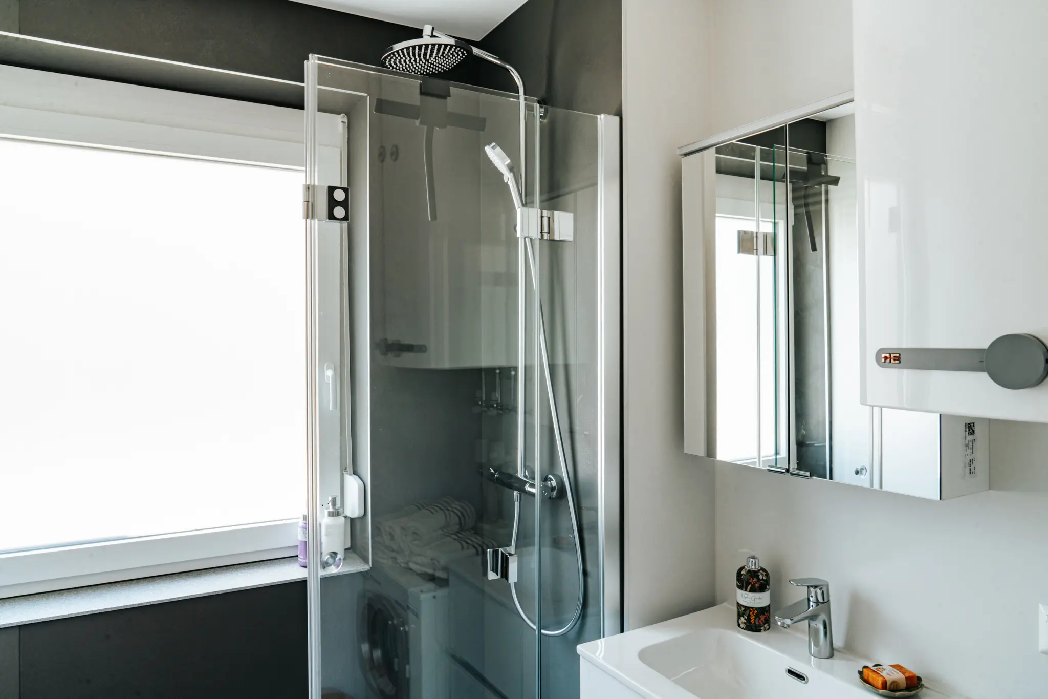 Ein modernes Badezimmer im Appartement Grand Prix in Zeltweg, ausgestattet mit einer geräumigen Glasdusche mit Regenduschkopf, einem Waschbecken mit integriertem Spiegel und stilvollen Details. Das helle Tageslicht, das durch das große Fenster fällt, schafft eine angenehme Atmosphäre und ergänzt die hochwertige Einrichtung perfekt. Ideal für Gäste, die nach einem langen Tag Entspannung suchen, sei es nach einem Besuch am Red Bull Ring oder einem Erkundungstag in der steirischen Umgebung.