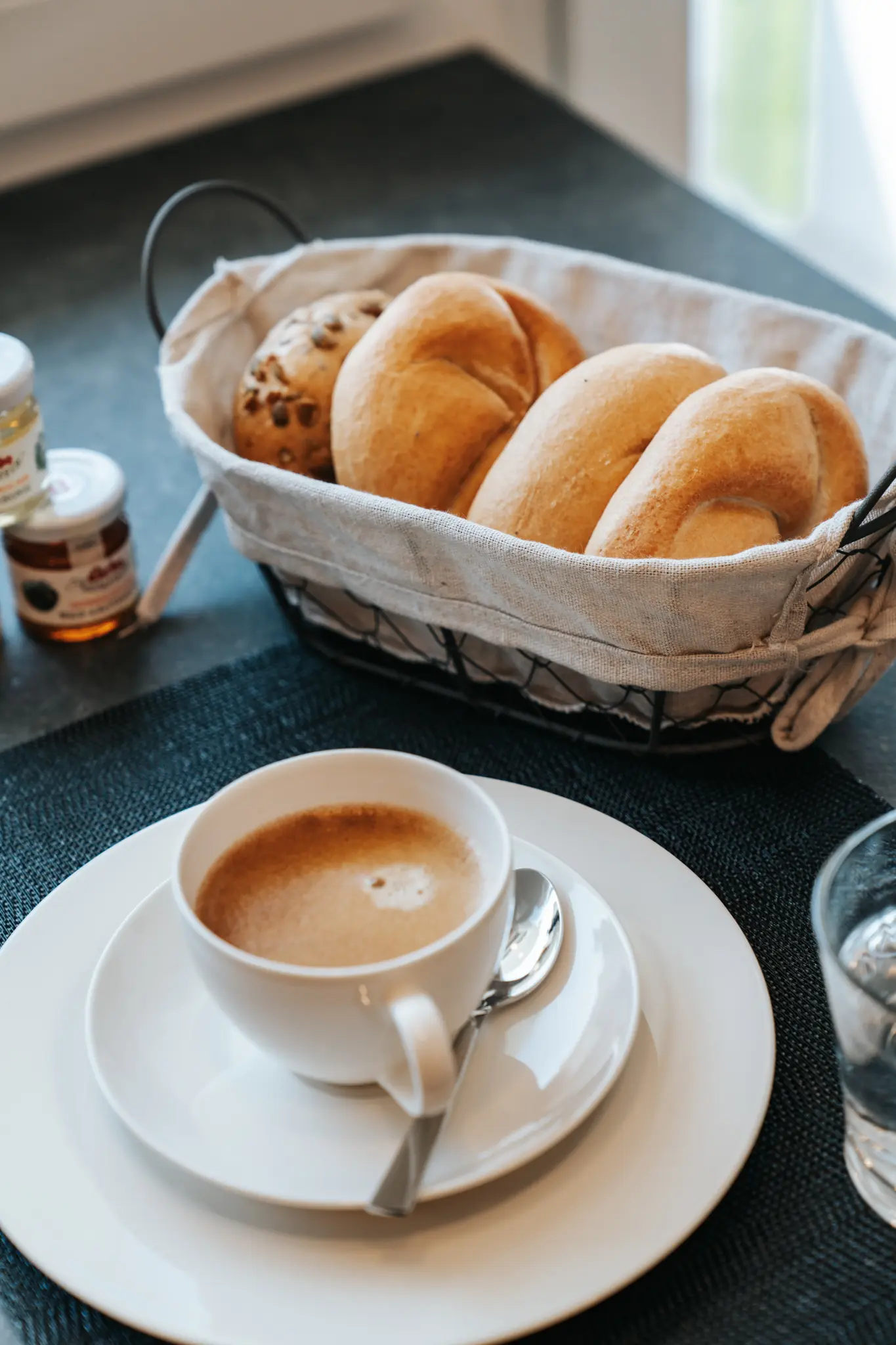 Frühstücksszene mit einer weißen Kaffeetasse, gefüllt mit einem Cappuccino, auf einem gedeckten Tisch. Daneben steht ein Brotkorb mit frischen Semmeln und einem Vollkornbrötchen sowie kleine Marmeladengläser und ein Glas Wasser.