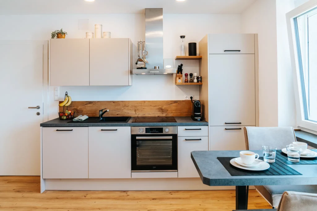 Eine moderne, stilvolle Küche im Appartement Grand Prix in Zeltweg, ausgestattet mit weißen Schränken, eleganten Holzdetails und hochwertigen Geräten wie einem Backofen, Herd und Kaffeemaschine. Perfekt für Gäste, die beim Formel 1-Rennen in Spielberg höchsten Komfort und die Möglichkeit zur Selbstverpflegung genießen möchten.