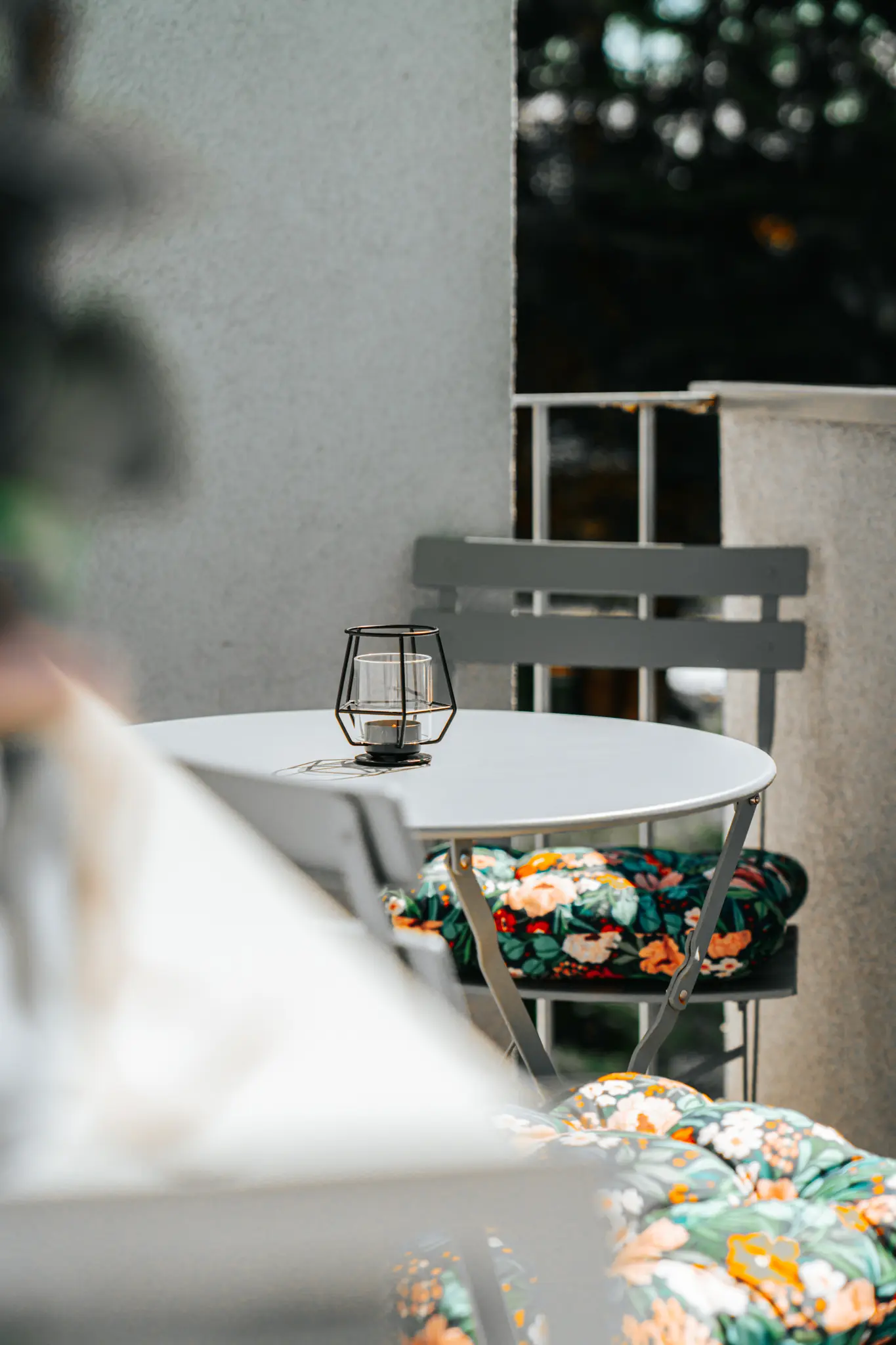 Ein stilvoll eingerichteter Balkon des luxuriösen Grand Prix Appartements in Zeltweg, nur wenige Minuten vom berühmten Red Bull Ring entfernt. Der Balkon bietet einen kleinen runden Tisch mit grauen Klappstühlen, die mit farbenfrohen Blumenkissen ausgestattet sind. Eine schlichte Laterne auf dem Tisch rundet die gemütliche Atmosphäre ab.