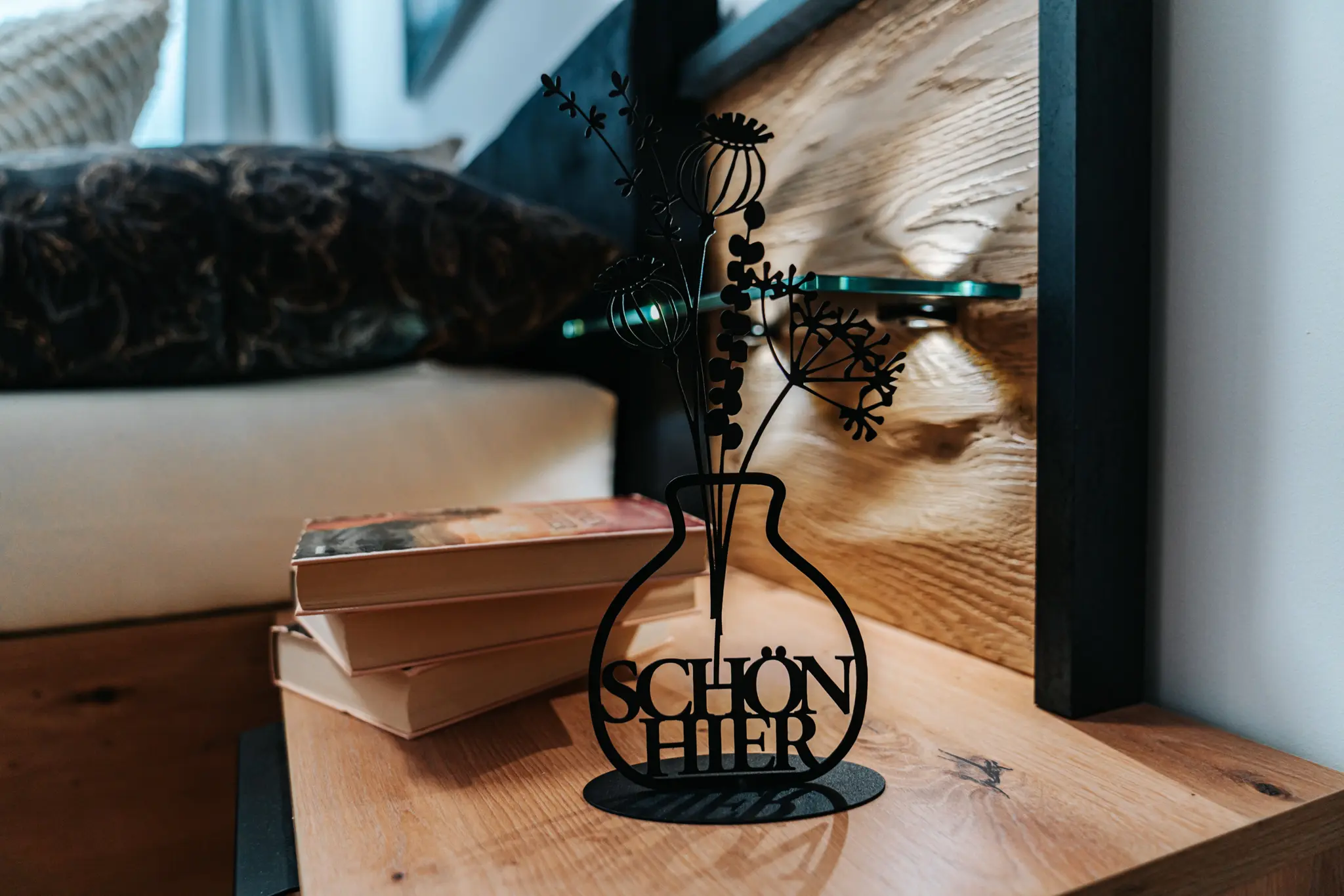 Dekoratives Detail im luxuriösen Grand Prix Appartement in Zeltweg: Eine stilvolle schwarze Metallvase mit filigranen Pflanzenmotiven und der Aufschrift "Schön hier", arrangiert auf einem Holznachttisch neben einem Stapel Bücher. Die Kombination aus natürlichen Materialien und moderner Dekoration verleiht dem Raum eine gemütliche und hochwertige Atmosphäre.