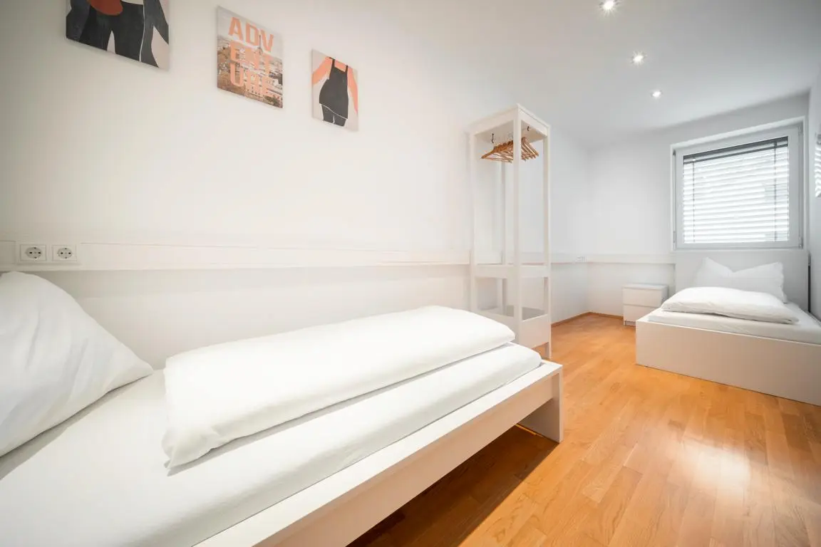 Das Bild zeigt ein helles und modernes Zweibettzimmer im CityFLAT 38 Appartement by globboard in Schladming-Dachstein. Mit klarer, minimalistischer Einrichtung und Holzboden bietet es eine komfortable Unterkunft für Freunde oder Familien.